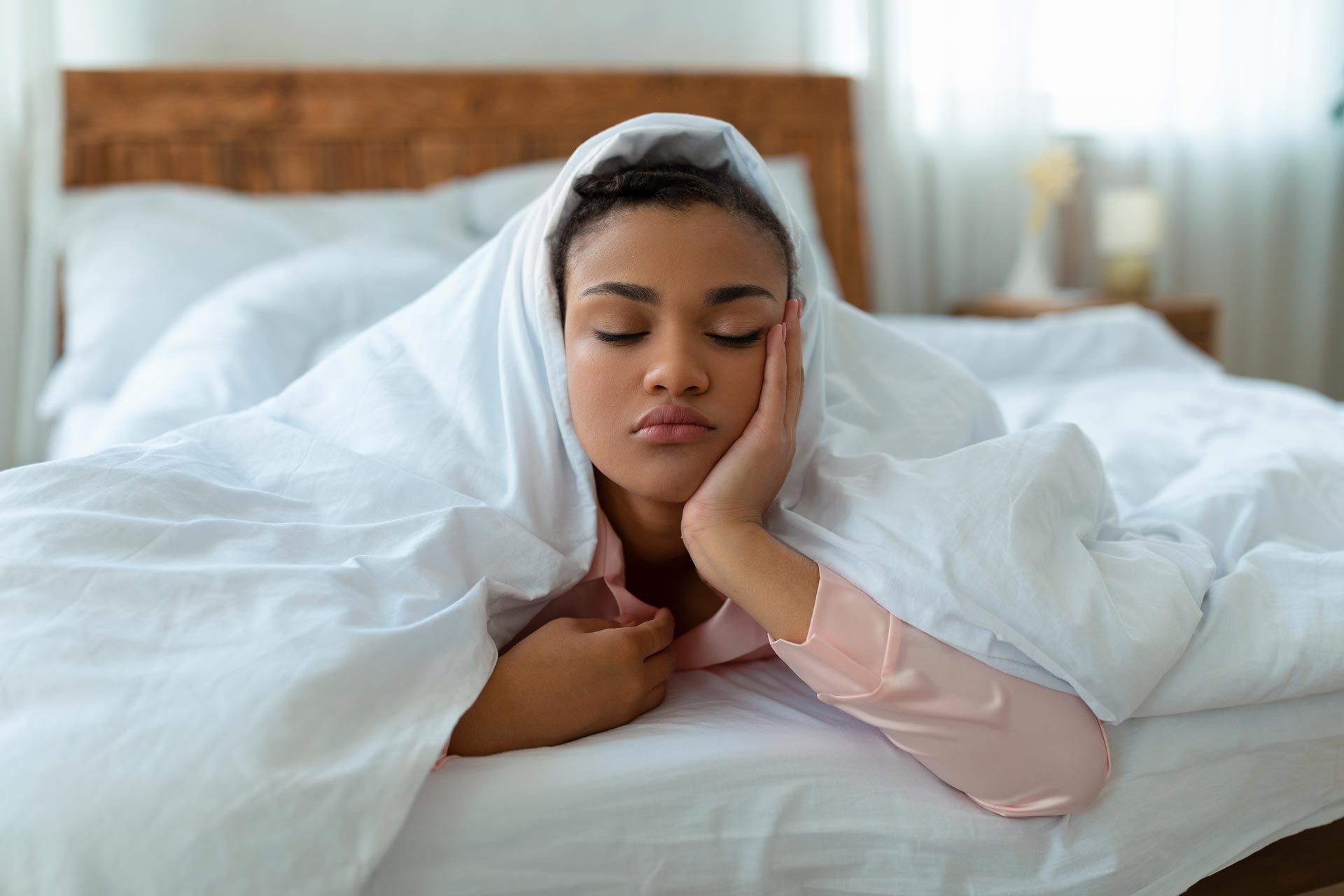 Person resting in bed with eyes closed, wrapped in white bedding and a blanket on a sunny morning