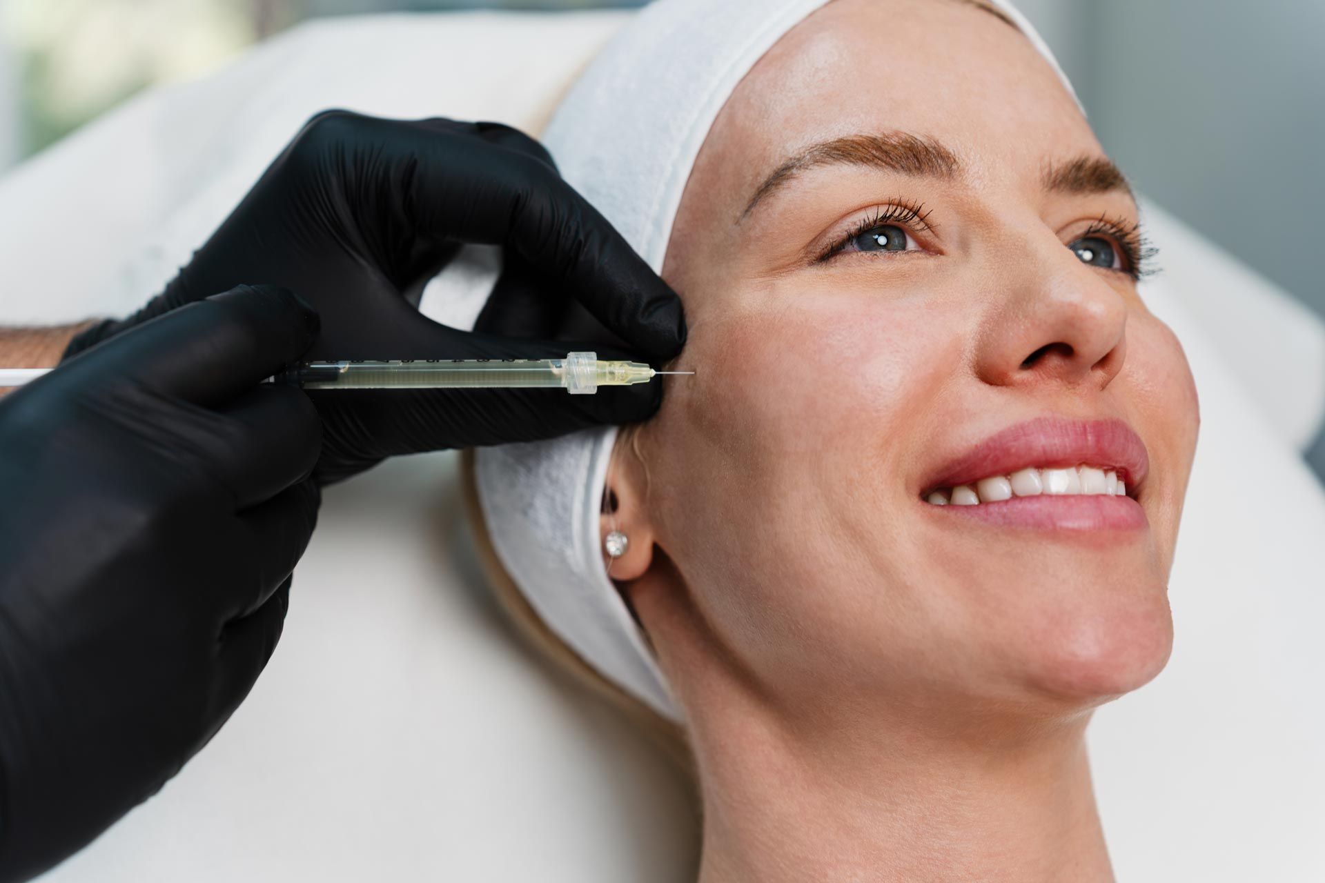 Gloved hand administering cosmetic injection to a smiling patient’s cheek