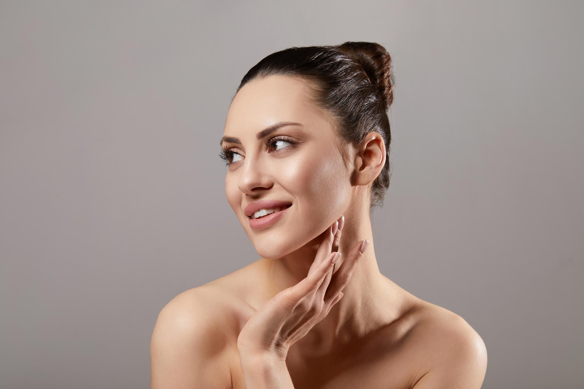 Smiling woman with dark hair in a bun touching her face against a gray background