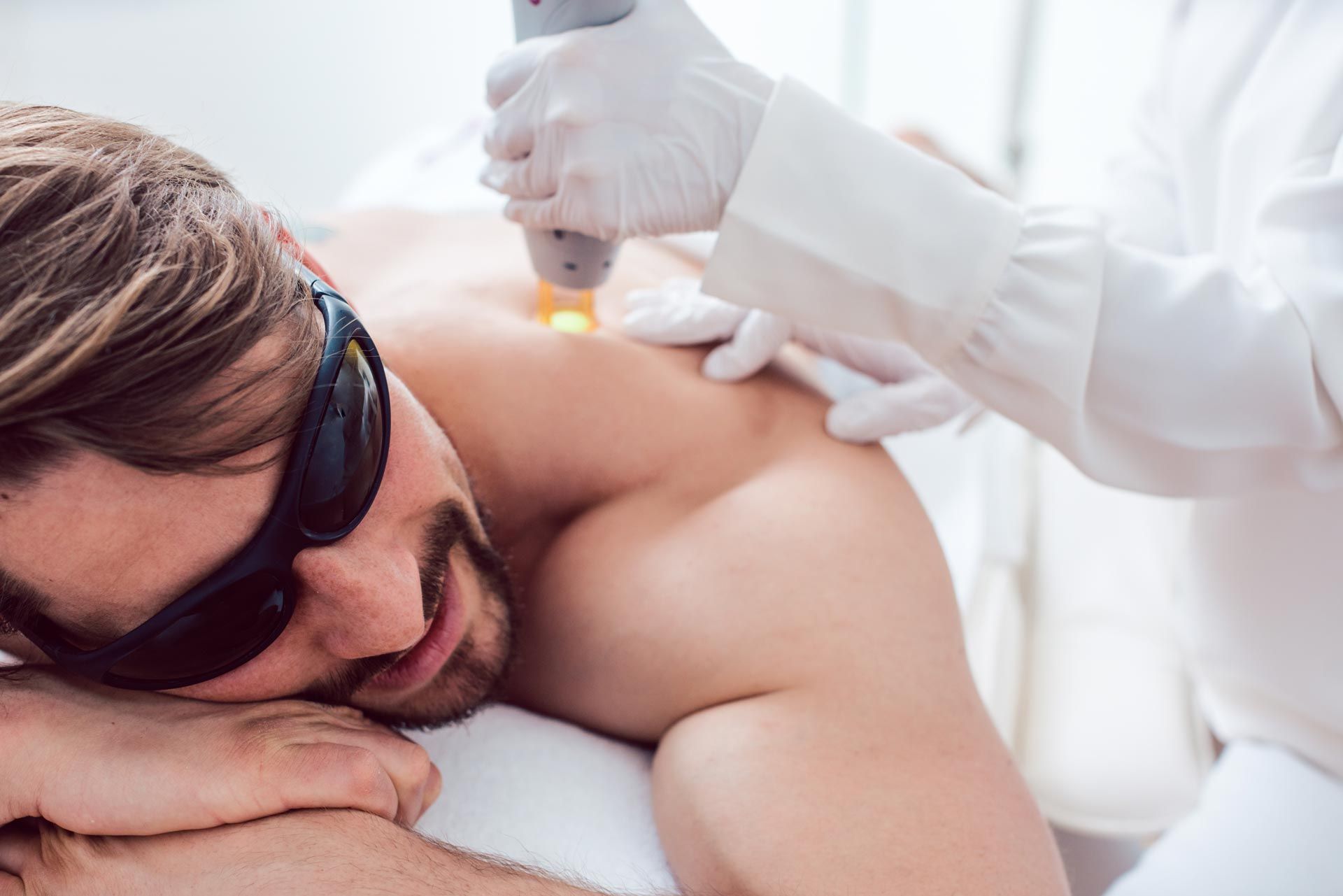 A man wearing protective goggles lies on a treatment bed as a gloved clinician applies a handheld device to his back.