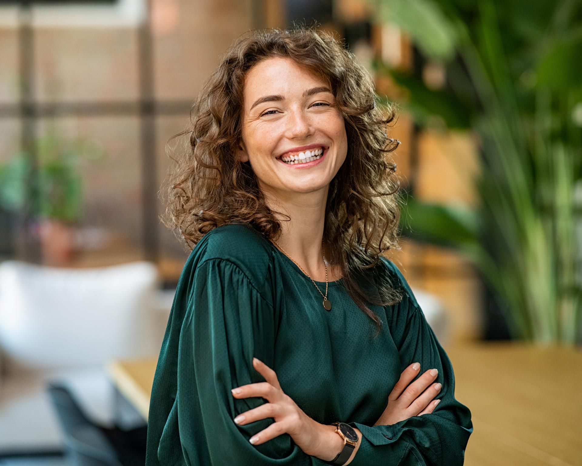 Smiling woman with curly hair, arms crossed, standing in a modern indoor office setting