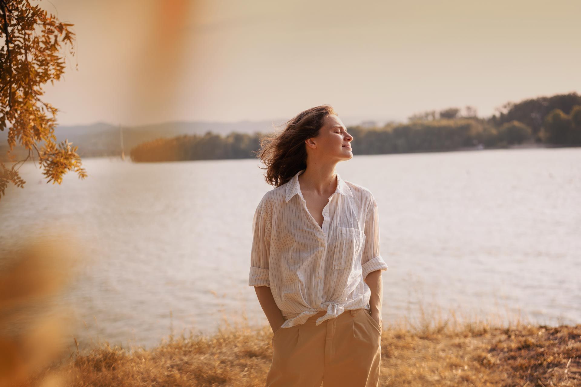 Person in a white shirt stands by a lake at sunset, looking upward in warm golden light.