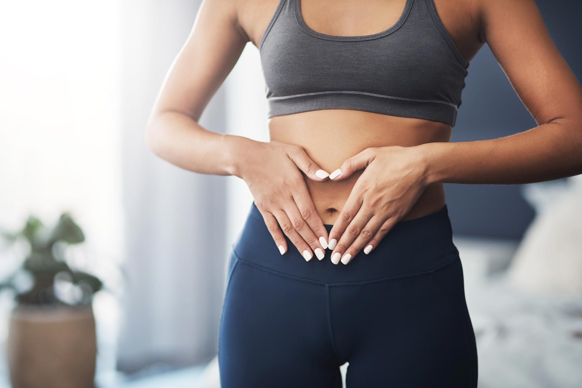 Person in gray sports bra and navy leggings forming a heart shape over their lower abdomen indoors.