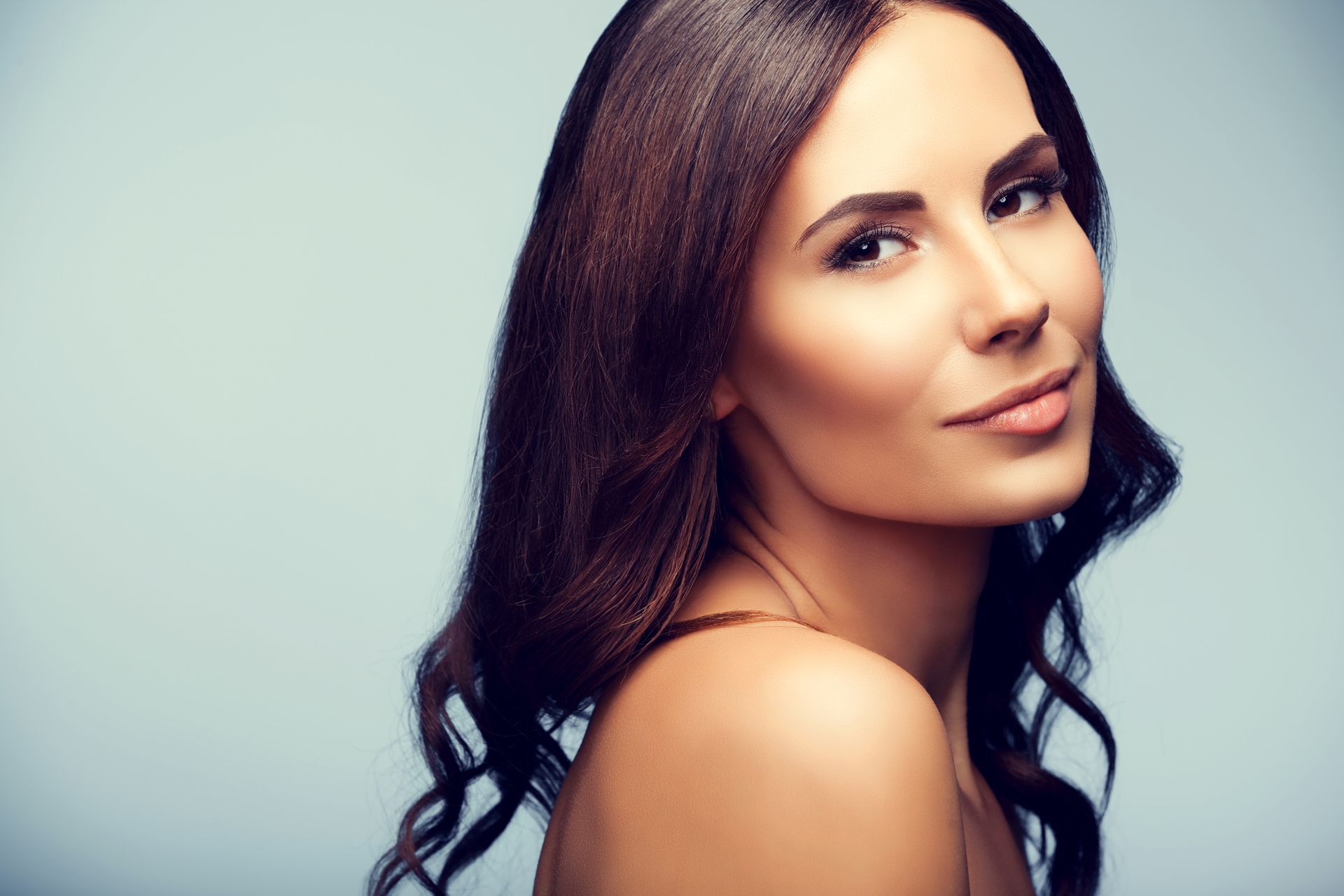 Woman smiling with cream under her eyes, gently touching her face against a light background