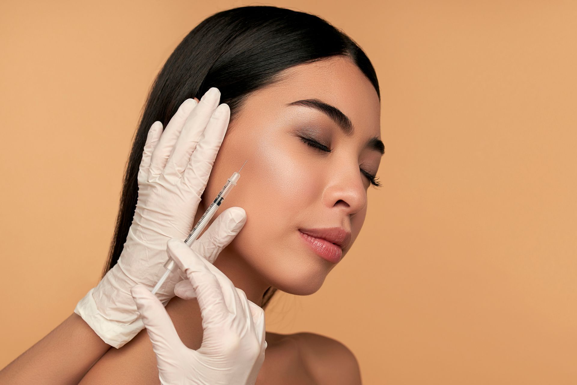 Gloved hands injecting filler into a woman’s cheek against a peach background