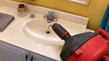 Red power drill aimed at a bathroom sink faucet with water running.