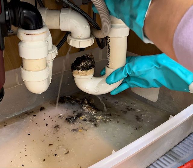 Gloved hands unclogging a dirty sink drain under white plumbing pipes, with wastewater in the basin