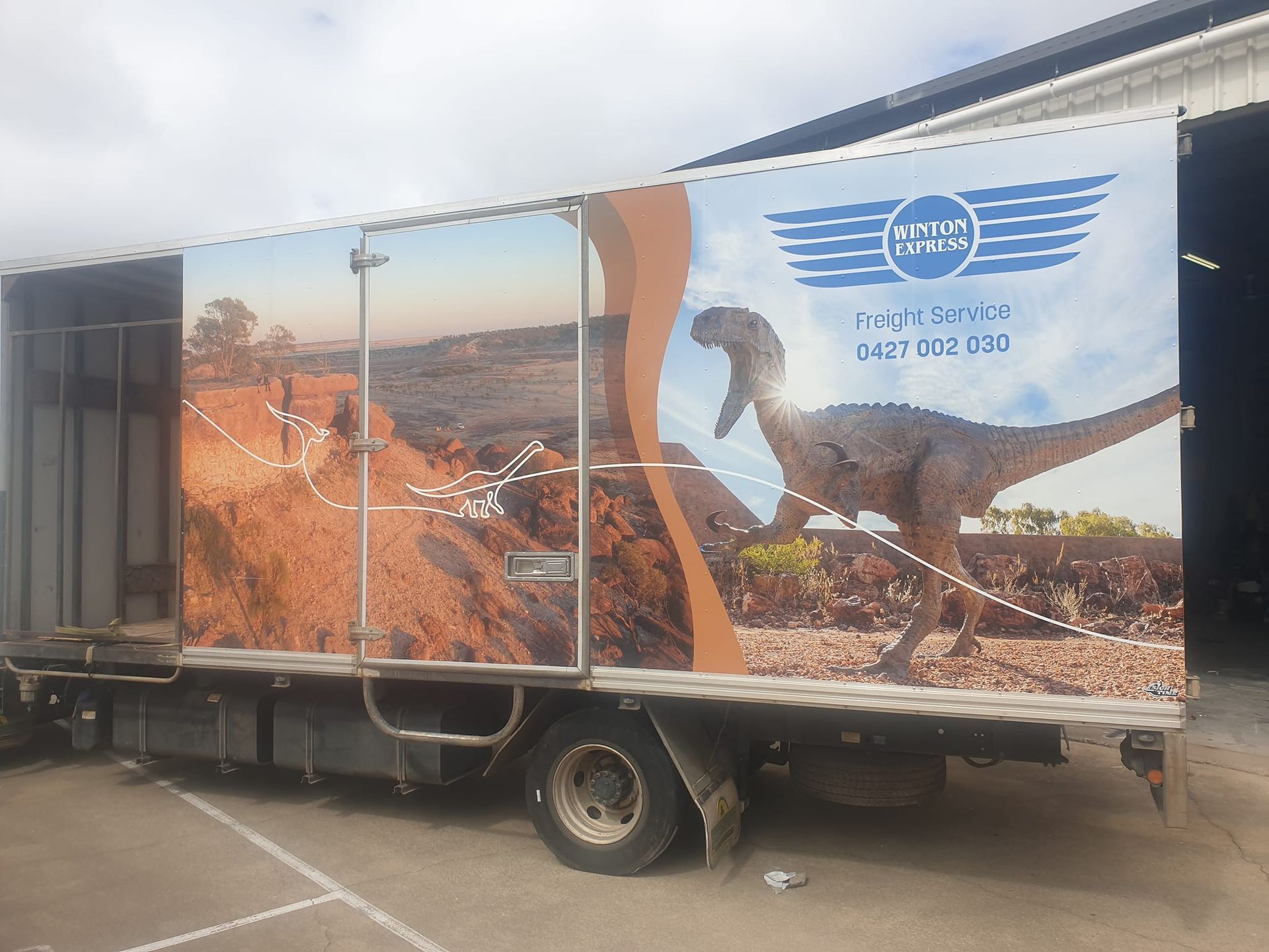 Truck with dinosaur and landscape graphics, branded with business logo and phone number.