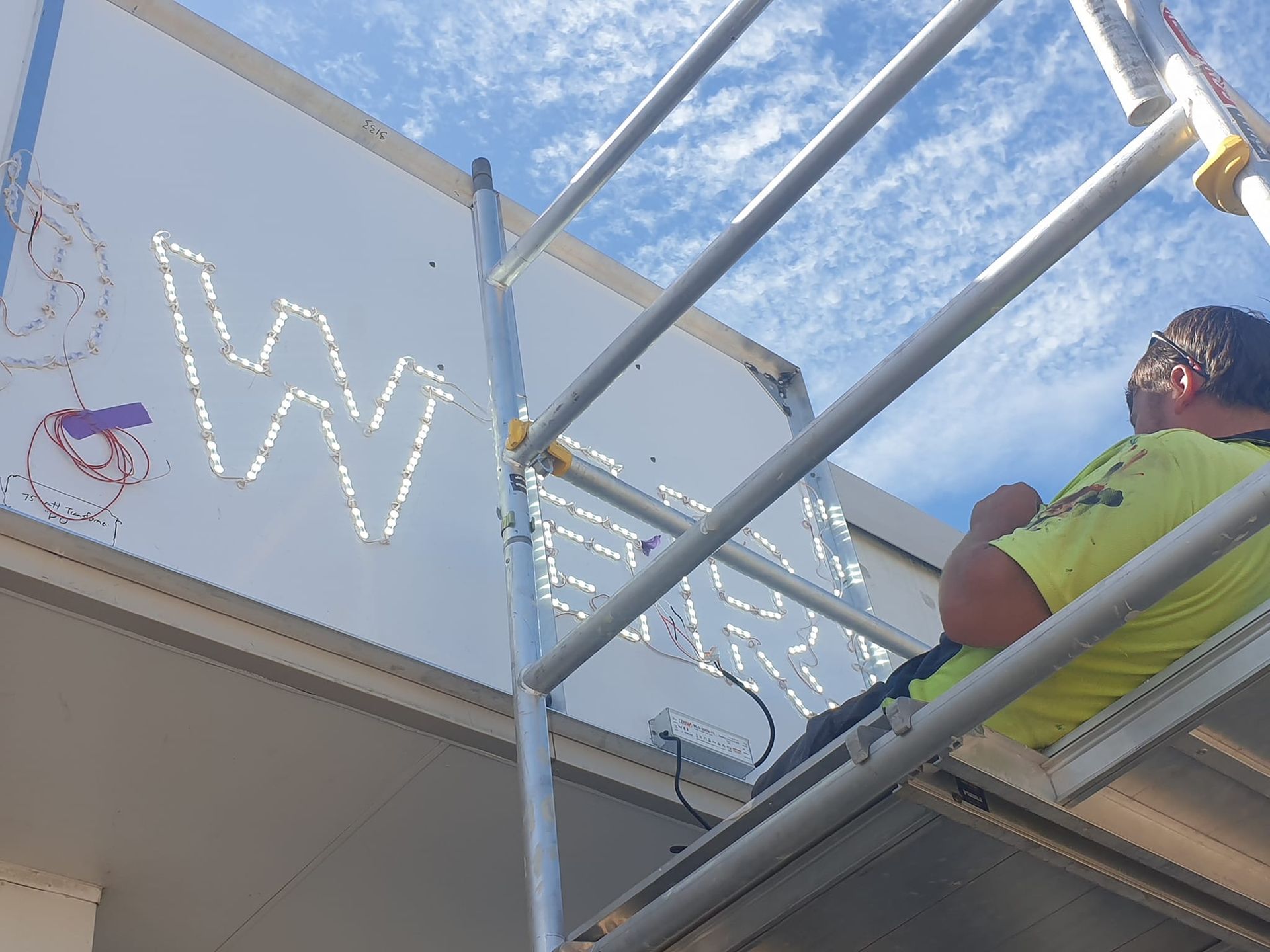 Man on scaffold installing a sign with illuminated letters against a partly cloudy sky.