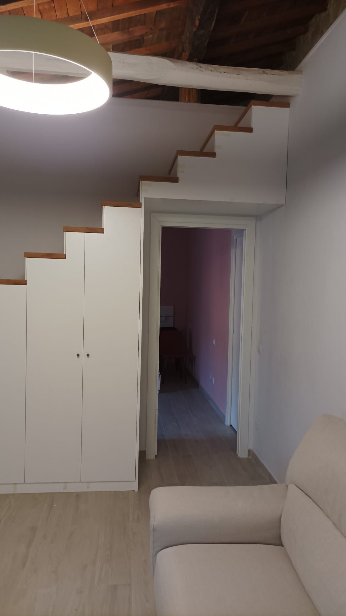 Piccola stanza con scala bianca, armadi a muro, porta e divano beige sotto un soffitto inclinato.