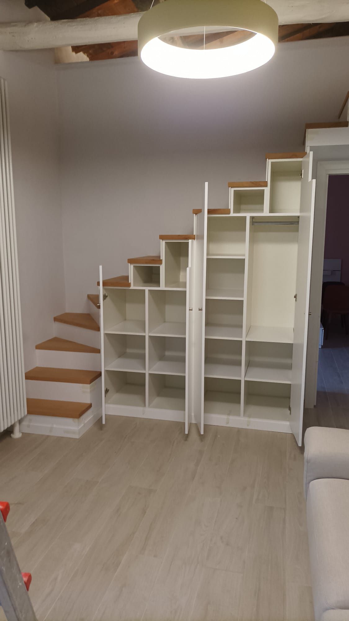 Interno della stanza con scaffalature bianche e una piccola scala in legno che conduce a una zona notte soppalcata.