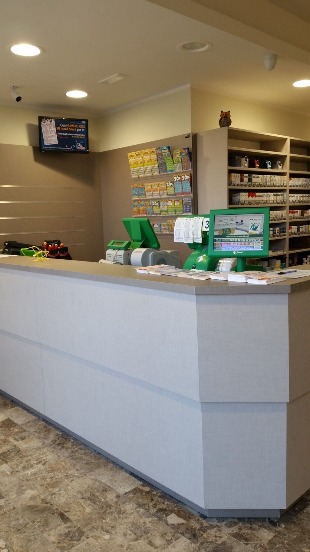 Bancone vuoto di una farmacia al dettaglio con insegne verdi, scaffali e un televisore a parete in un interno luminoso del negozio.
