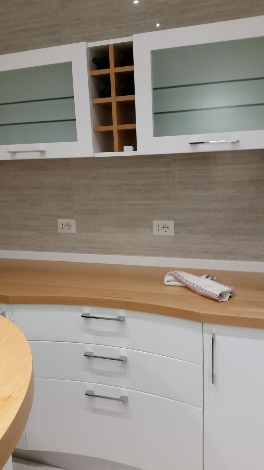 Cucina moderna con mobili bianchi, piano di lavoro in legno, paraschizzi piastrellato e pensili con ante in vetro.