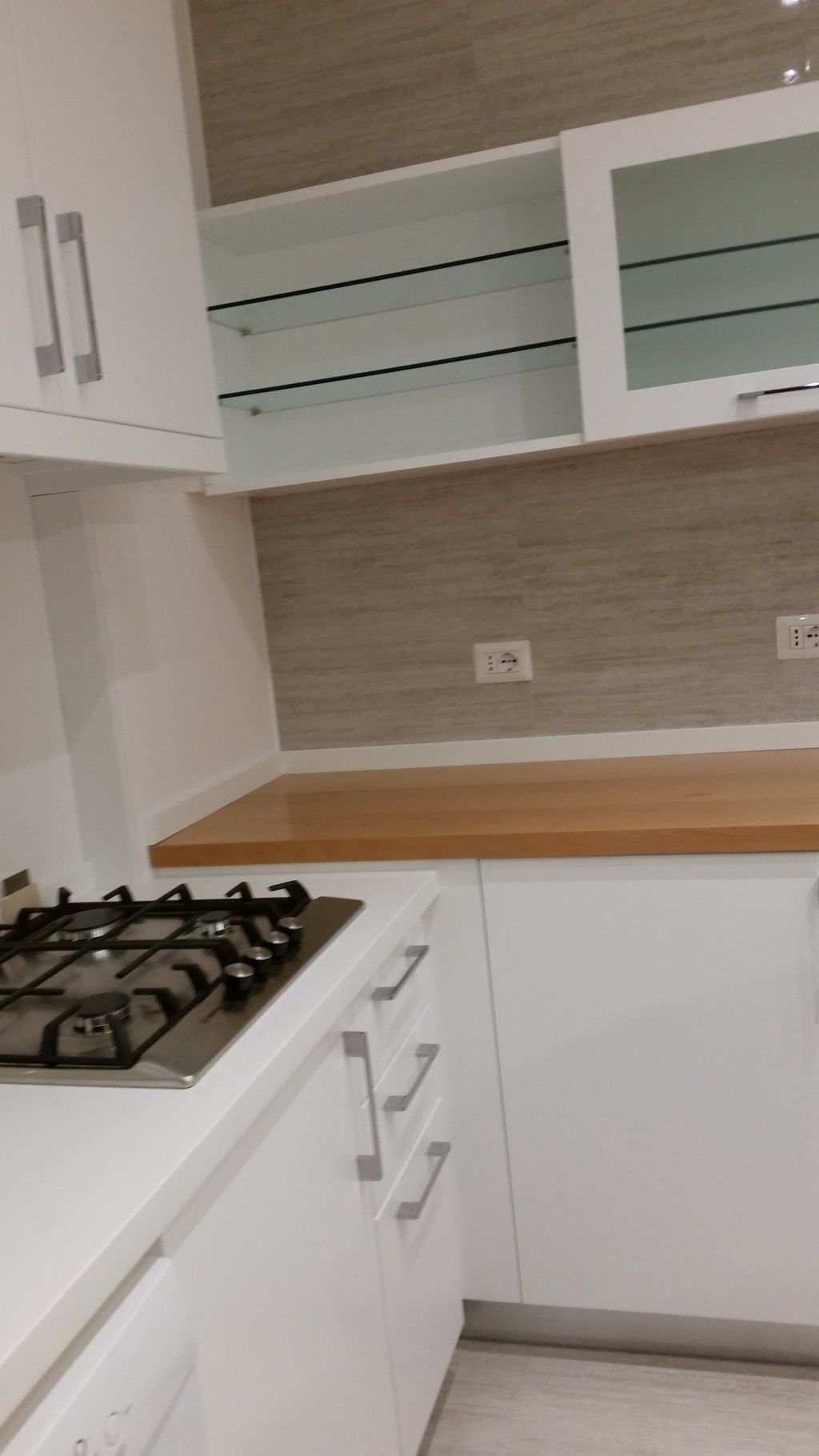 Cucina moderna bianca con piano cottura a gas, ripiano in legno e pensili con ante in vetro.