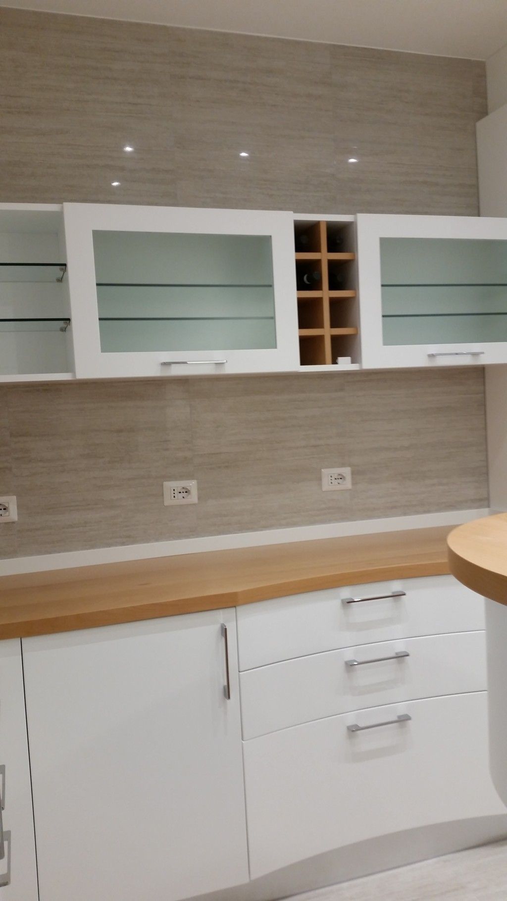 Parete cucina moderna con mobili bianchi, piano di lavoro in legno, paraschizzi beige e portabottiglie integrato.