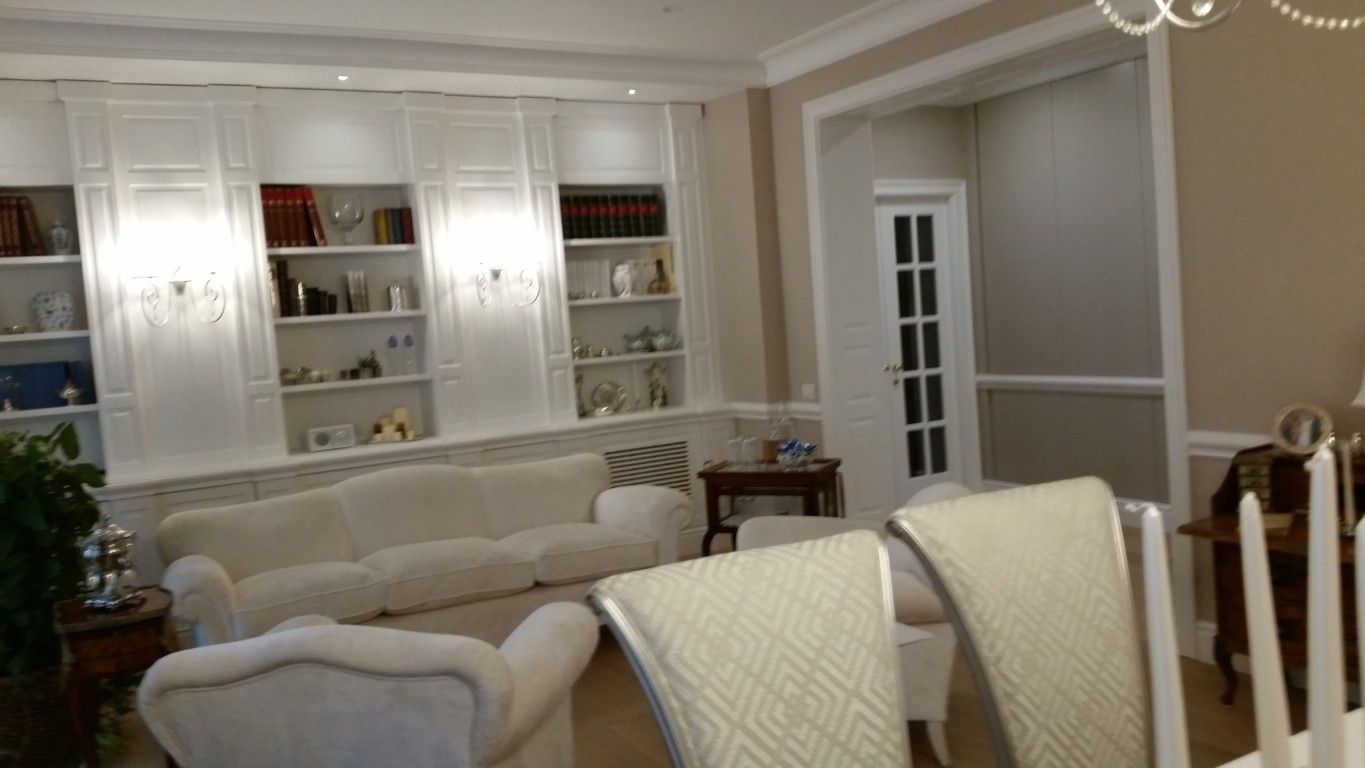 Elegante soggiorno bianco con scaffali a muro, mobili color crema e porta con pannelli in vetro.