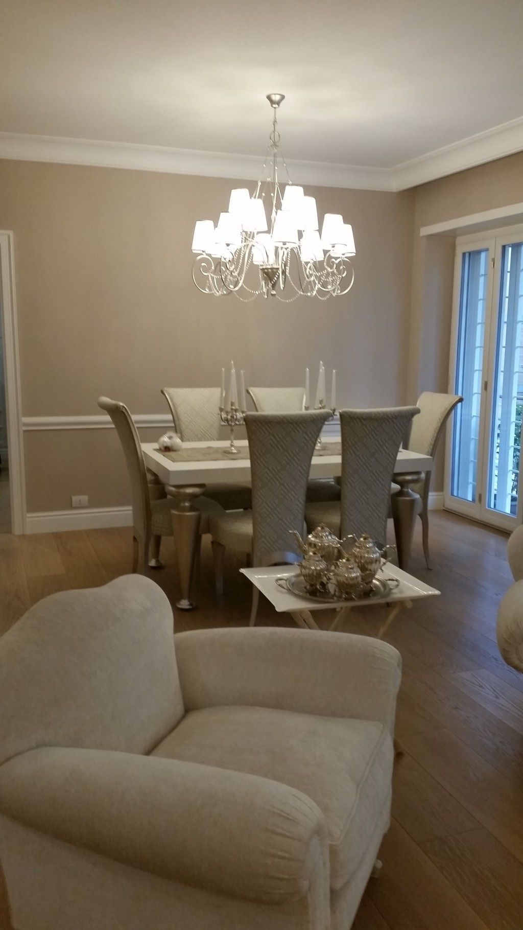 Elegante sala da pranzo con lampadario, sedie bianche e poltrona beige in primo piano.
