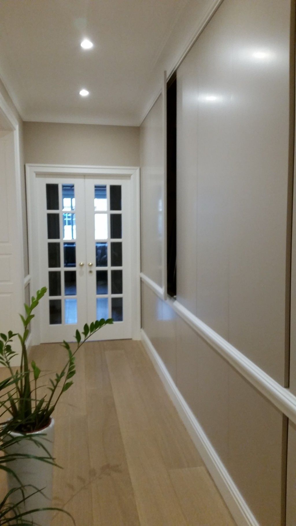 Stretto corridoio con pareti beige, faretti a incasso sul soffitto, finiture bianche e porte con pannelli di vetro in fondo.
