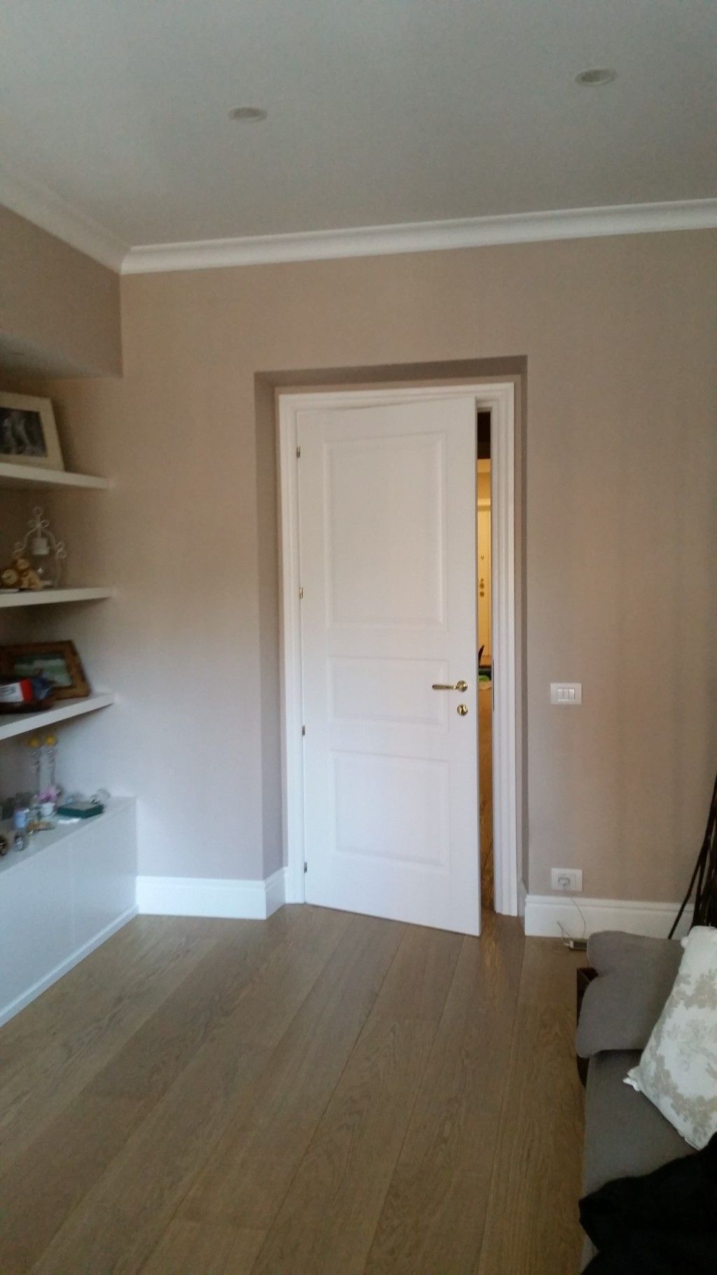 Piccola stanza beige con porta bianca, scaffali a muro sulla sinistra e sponda del letto sulla destra.