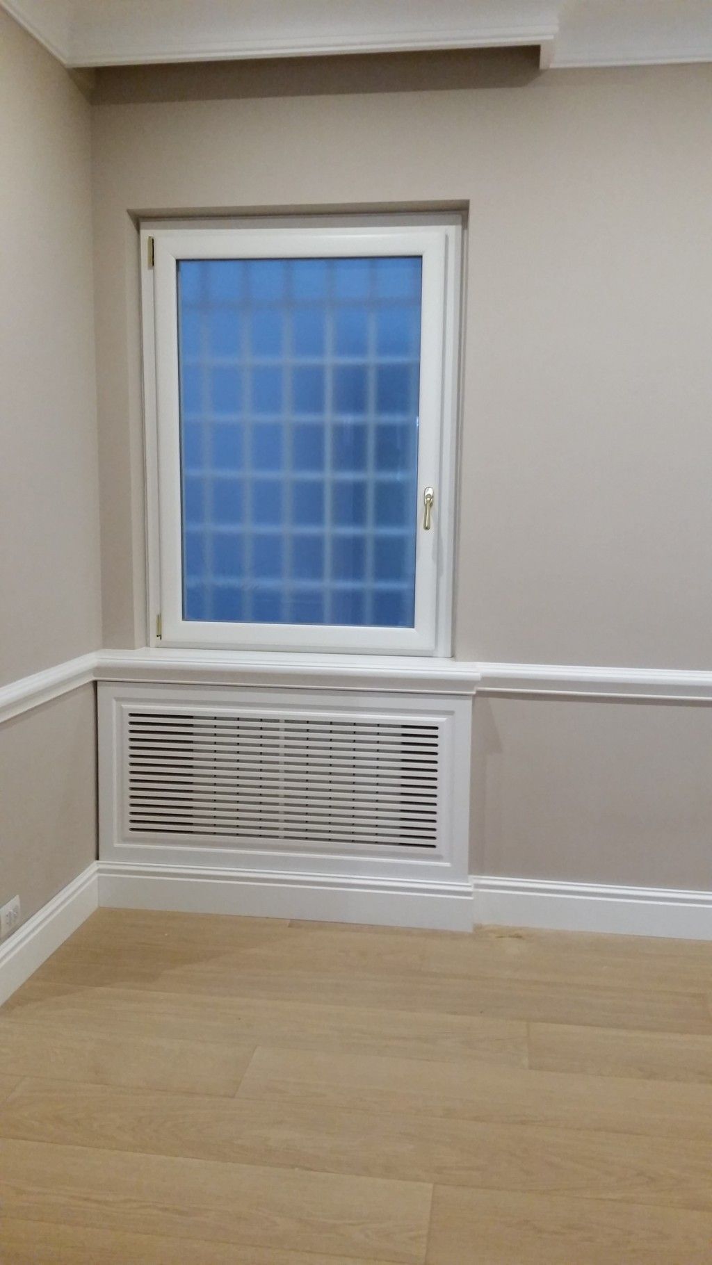 Piccola stanza vuota con finestra, pellicola oscurante blu, bocchetta di ventilazione a parete, pareti beige e pavimento in legno chiaro.
