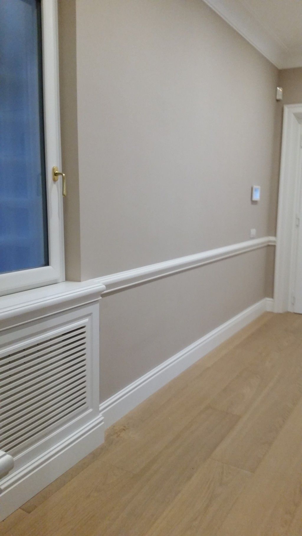 Corridoio vuoto color beige con finiture bianche, copritermosifone e pavimento in legno accanto a una finestra e una porta.