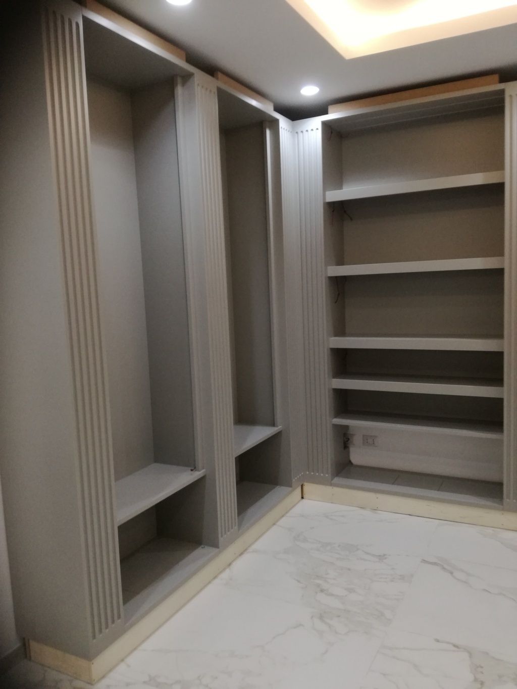 Cabina armadio a muro con ripiani beige, spazi per appendere gli abiti e pavimento piastrellato effetto marmo bianco.