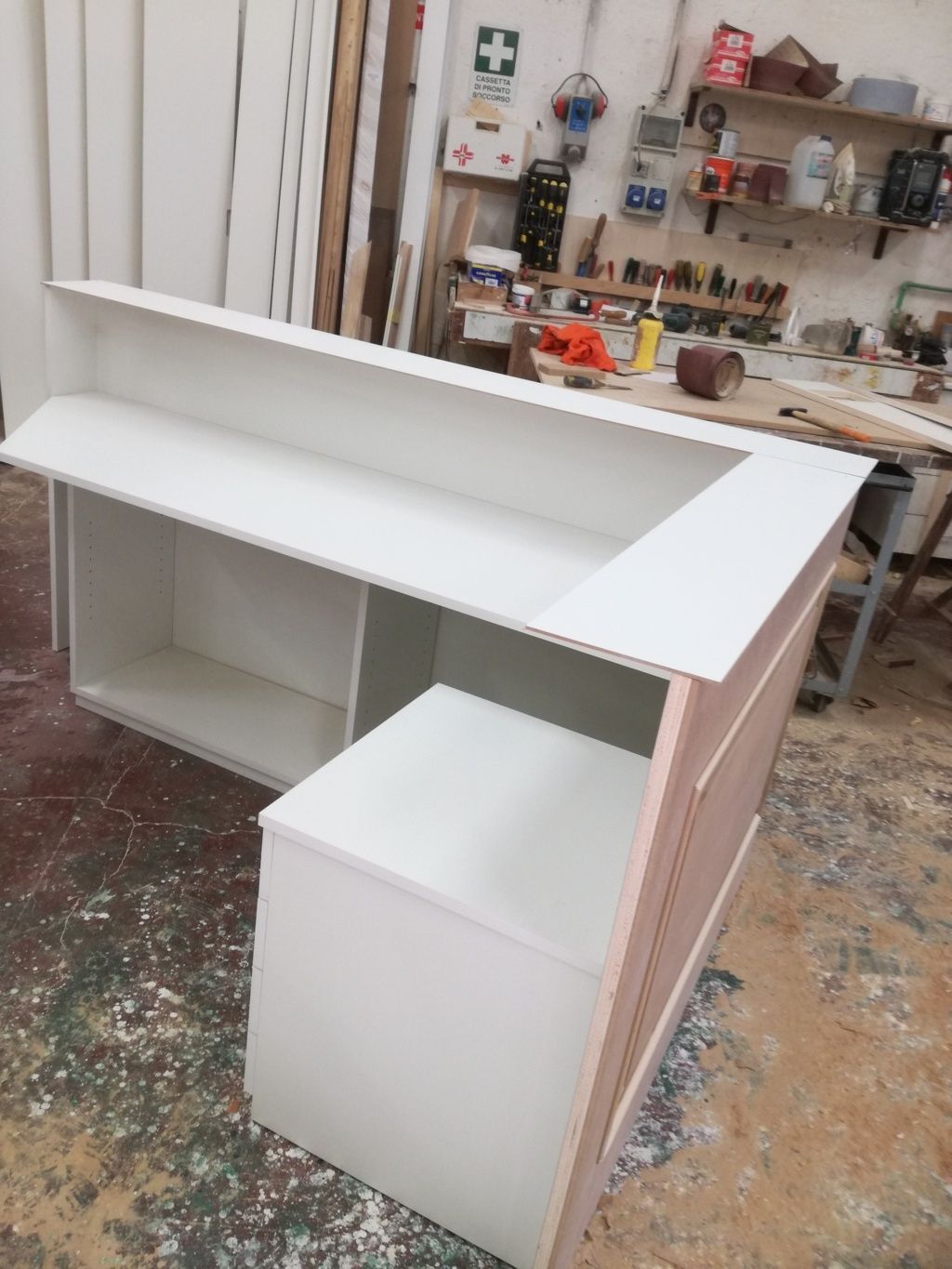 Bancone o scrivania a L di colore bianco in fase di costruzione in un'officina, con ripiani non rifiniti e piano inclinato.