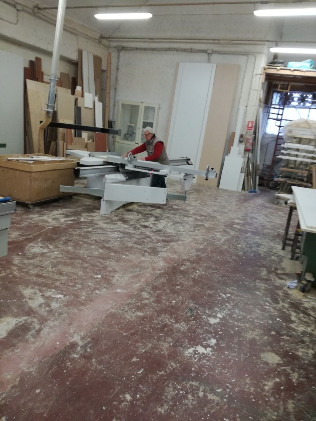 Laboratorio di falegnameria con pavimento ricoperto di segatura, tavoli da lavoro, attrezzi e una persona seduta vicino a un banco sega.