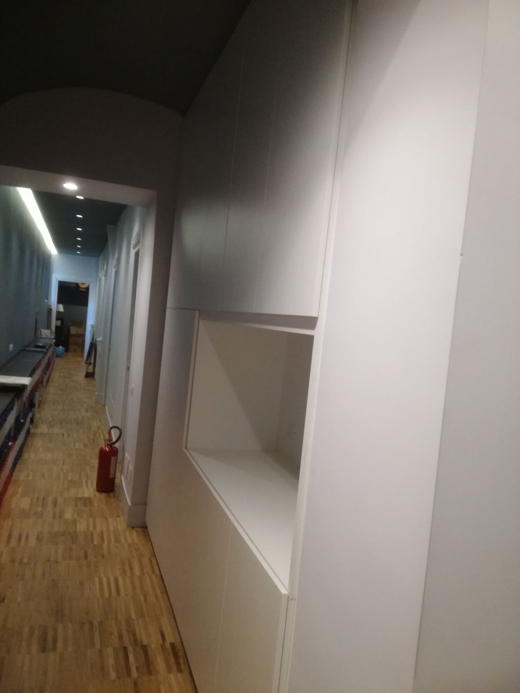 Stretto corridoio con pareti bianche, pavimento in legno e un estintore rosso in fondo.