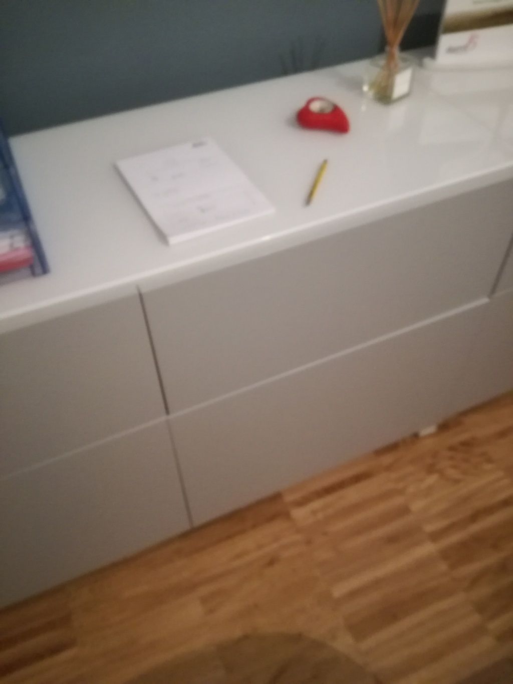 Un armadietto bianco con sopra un quaderno, una matita gialla e un metro a nastro rosso, accanto a un pavimento di legno.