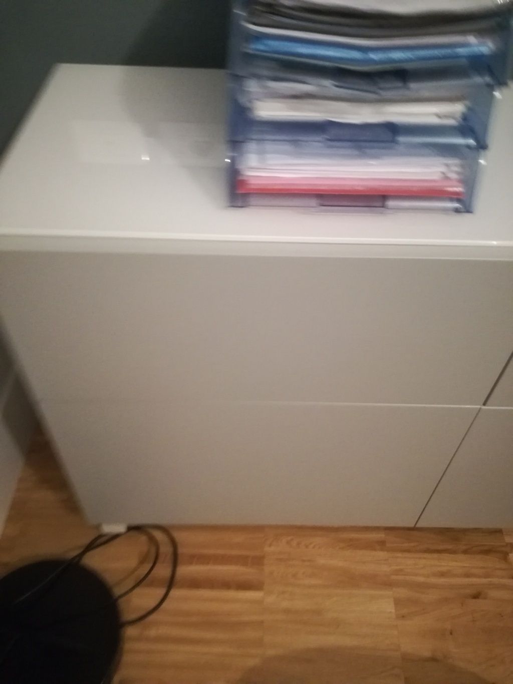 Mobile bianco con pile di carte e cartelle sulla parte superiore, accanto a un pavimento in legno e cavi sulla sinistra.
