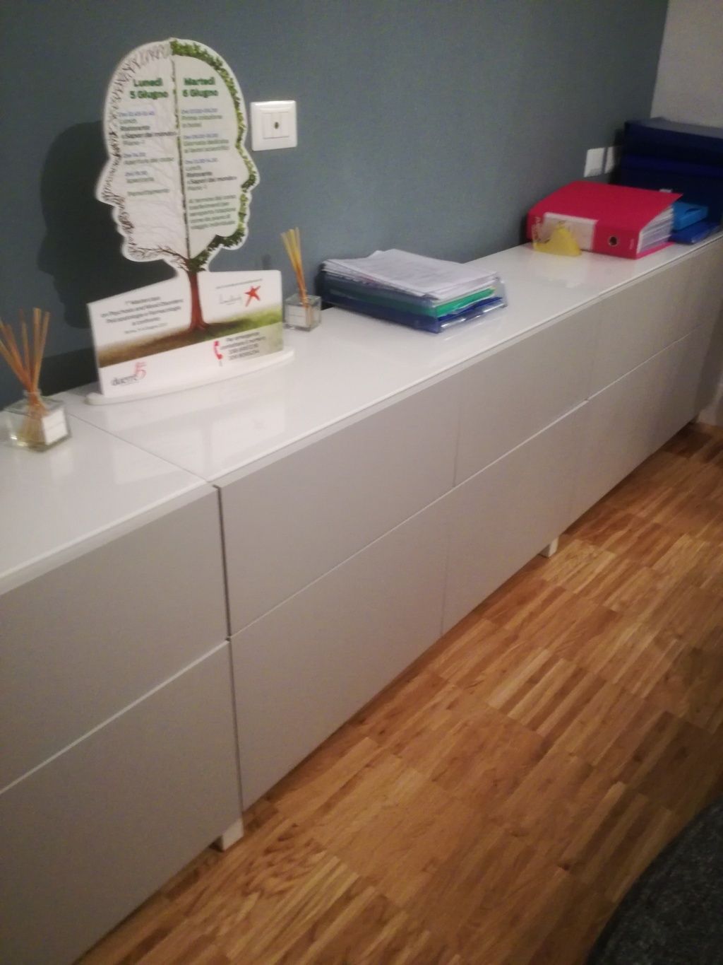 Lungo armadio bianco con documenti, un alberello decorativo e articoli di cancelleria, appoggiato a una parete grigia.