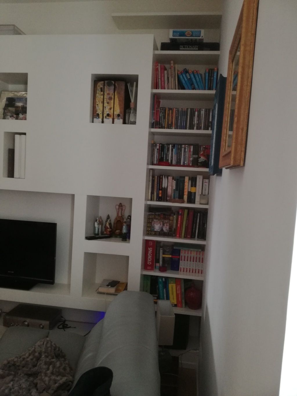 Soggiorno con scaffali bianchi, TV, libri, oggetti decorativi e una poltrona grigia in primo piano.