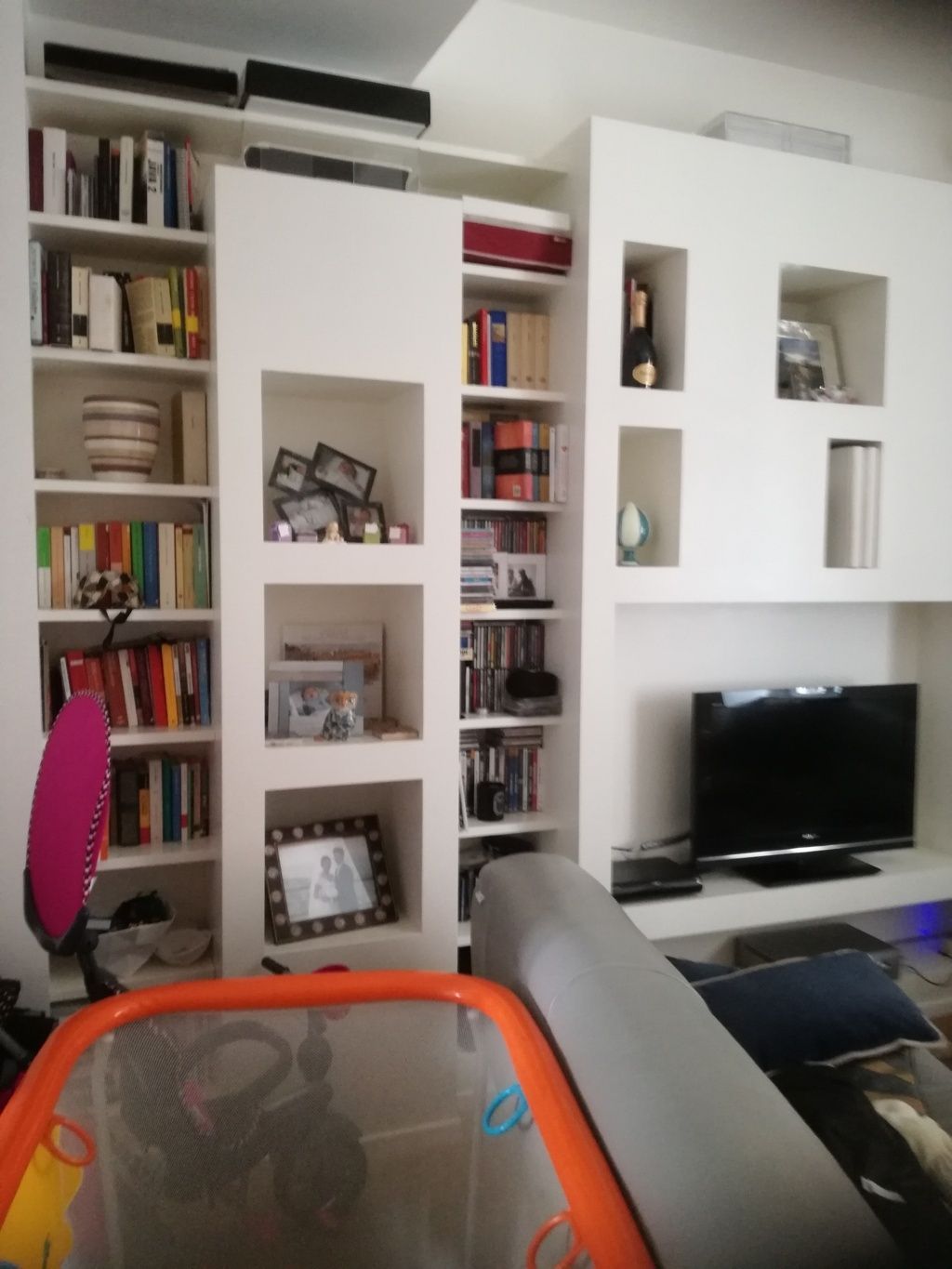 Soggiorno con scaffali a muro bianchi, TV, divano e, in primo piano, un box per bambini con cornice arancione.