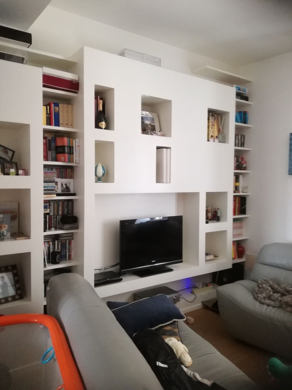 Soggiorno moderno con scaffali a muro bianchi, televisore e poltrona arancione.