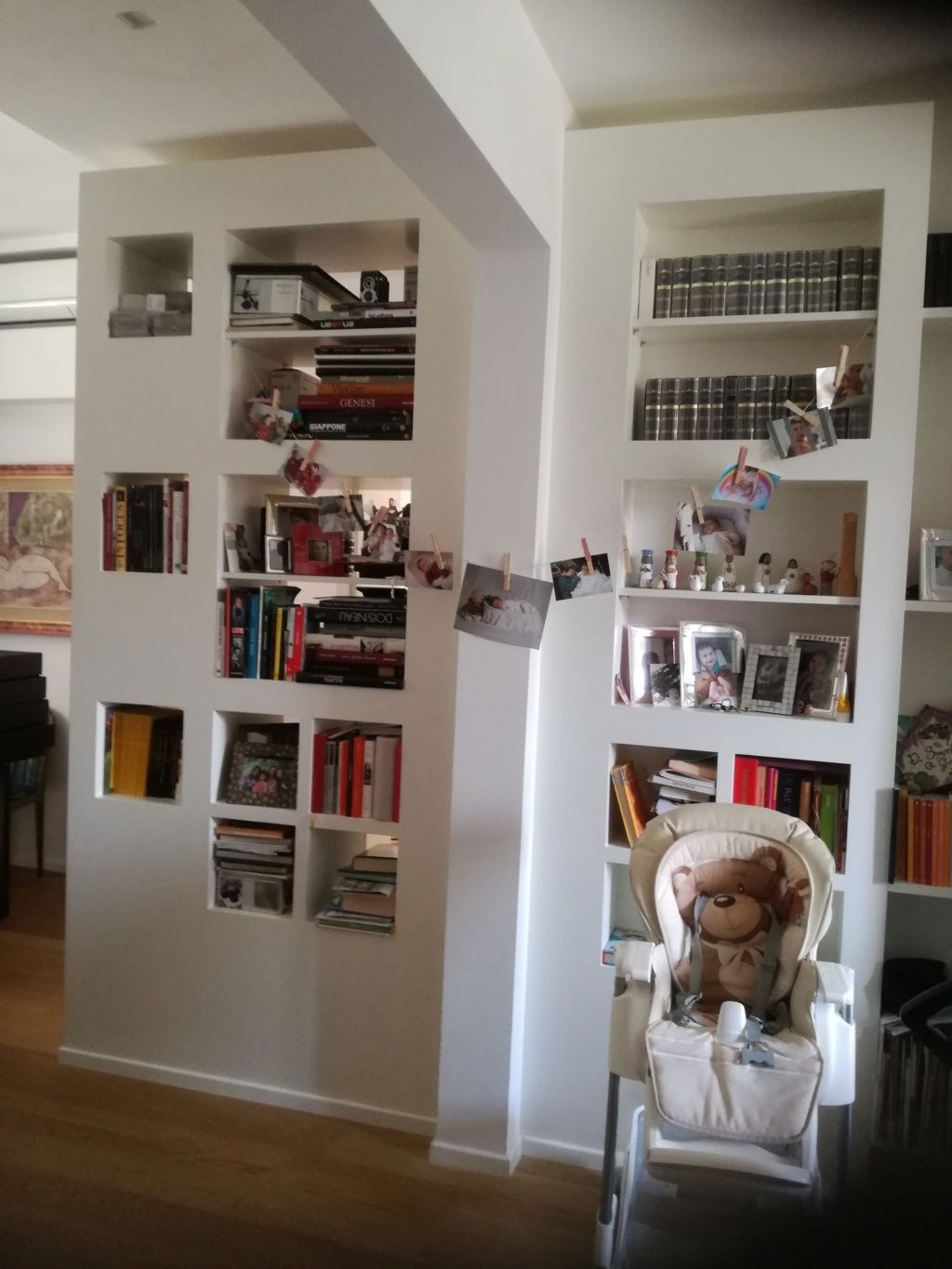 In una stanza luminosa, librerie a muro bianche con libri e oggetti decorativi; di fronte è posizionata un'altalena per bambini.