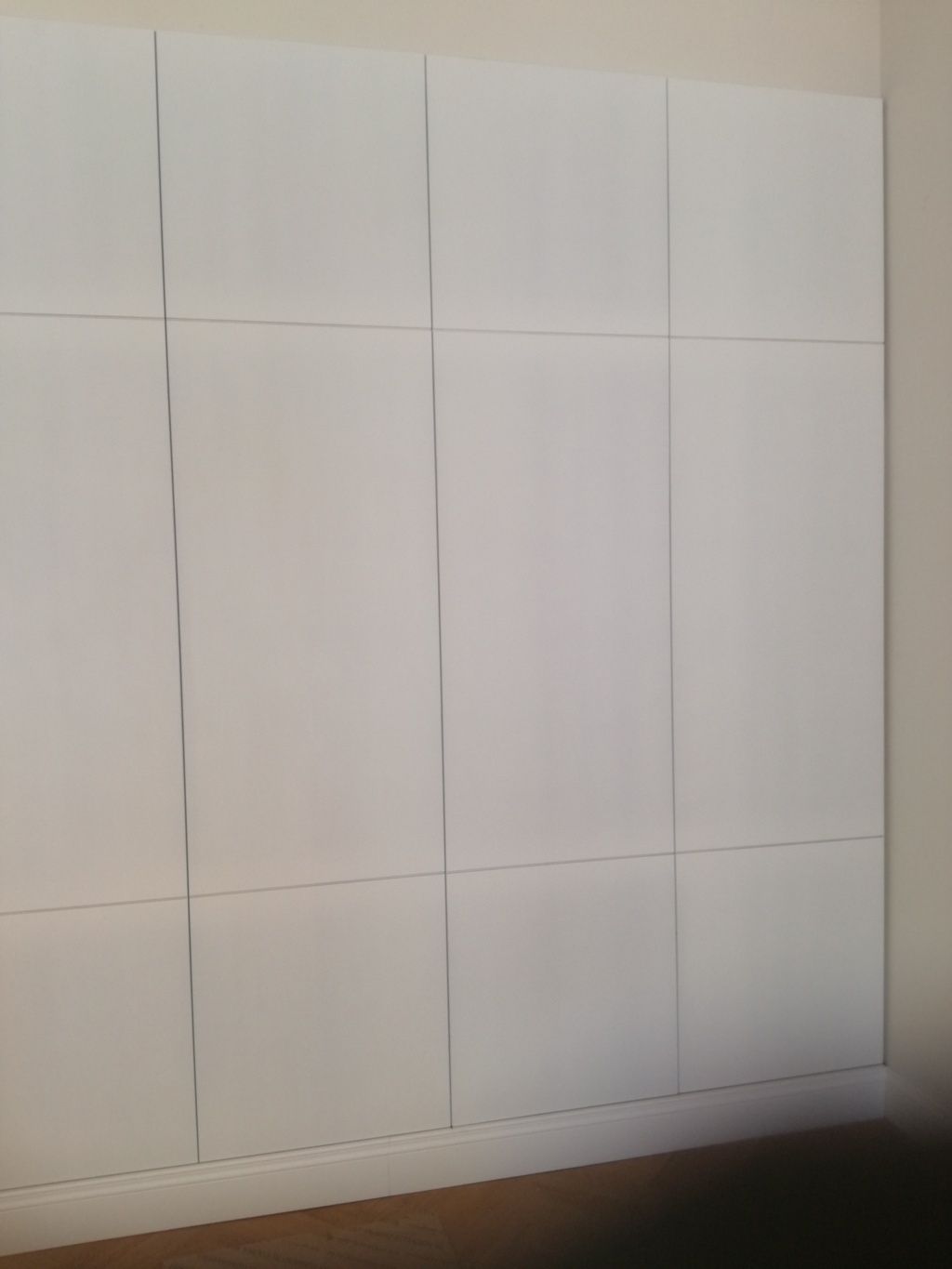 Parete rivestita di pannelli bianchi con sottili linee verticali e orizzontali sopra un pavimento in legno.