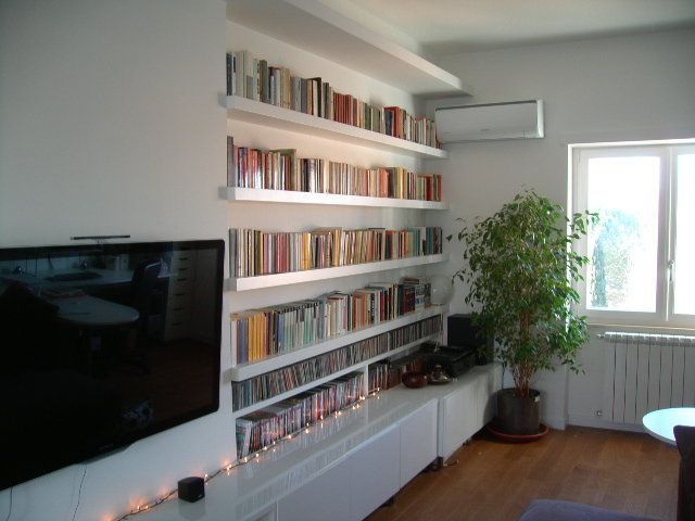Soggiorno con librerie a muro, TV, mobili bianchi e un'ampia finestra accanto a una pianta in vaso.