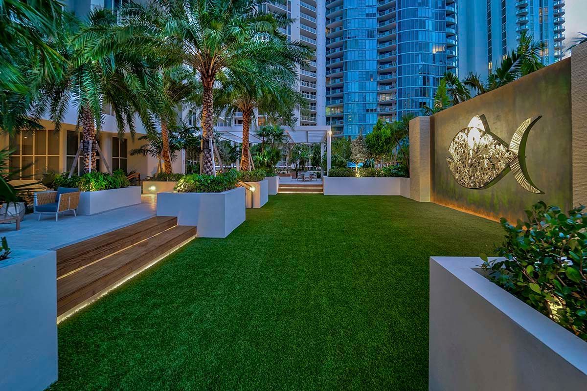 Modern apartment courtyard with green lawn, palm trees, planters, and city towers at dusk