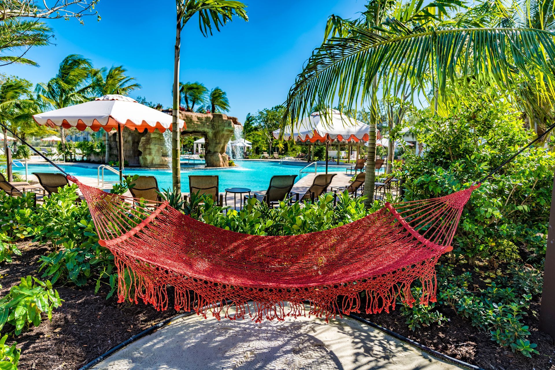 Red hammock in tropical garden beside a turquoise resort pool, with palm trees and white umbrellas.