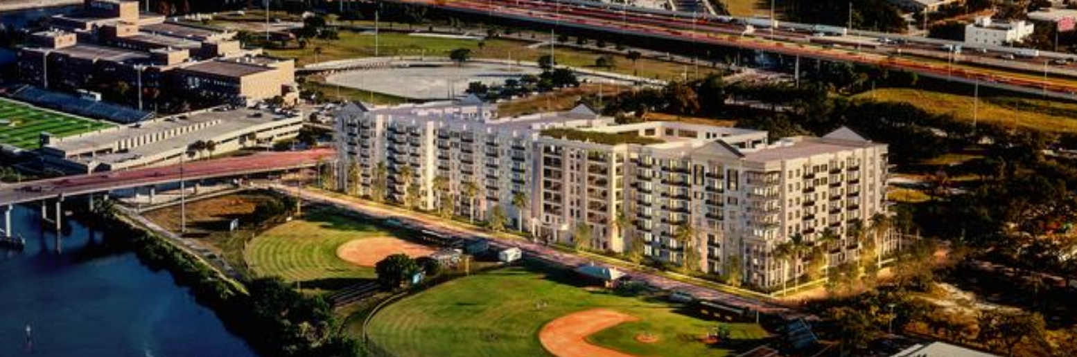 Aerial view of a riverside apartment complex beside a baseball field at sunset.