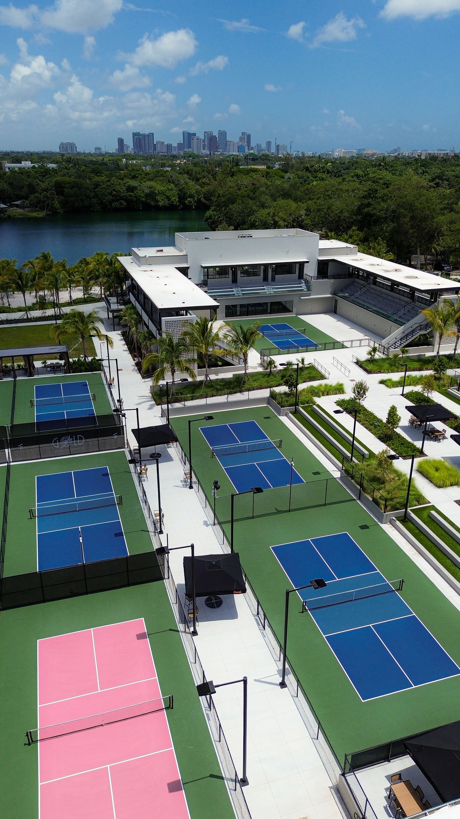 Aerial view of a riverside sports complex with multiple blue tennis courts and a central building