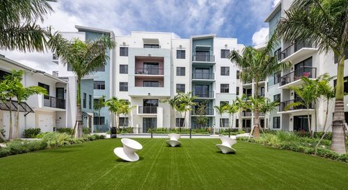 Modern white apartment complex with green lawn, palm trees, and courtyard seating under a blue sky