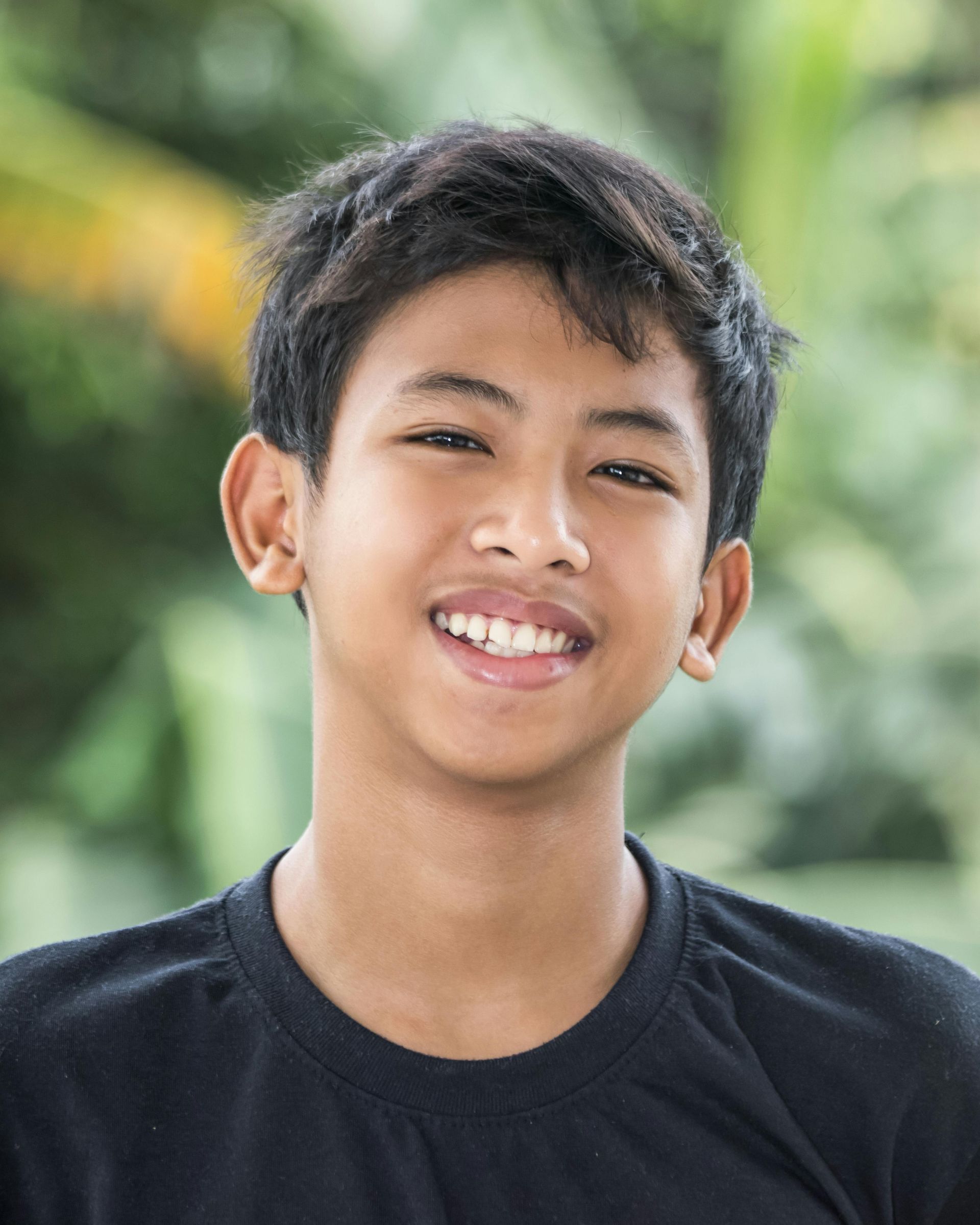 Smiling person with dark hair in a black shirt, outdoors with green foliage in the background.