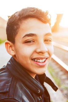 Boy smiling outdoors, wearing a black jacket, near train tracks at sunset.