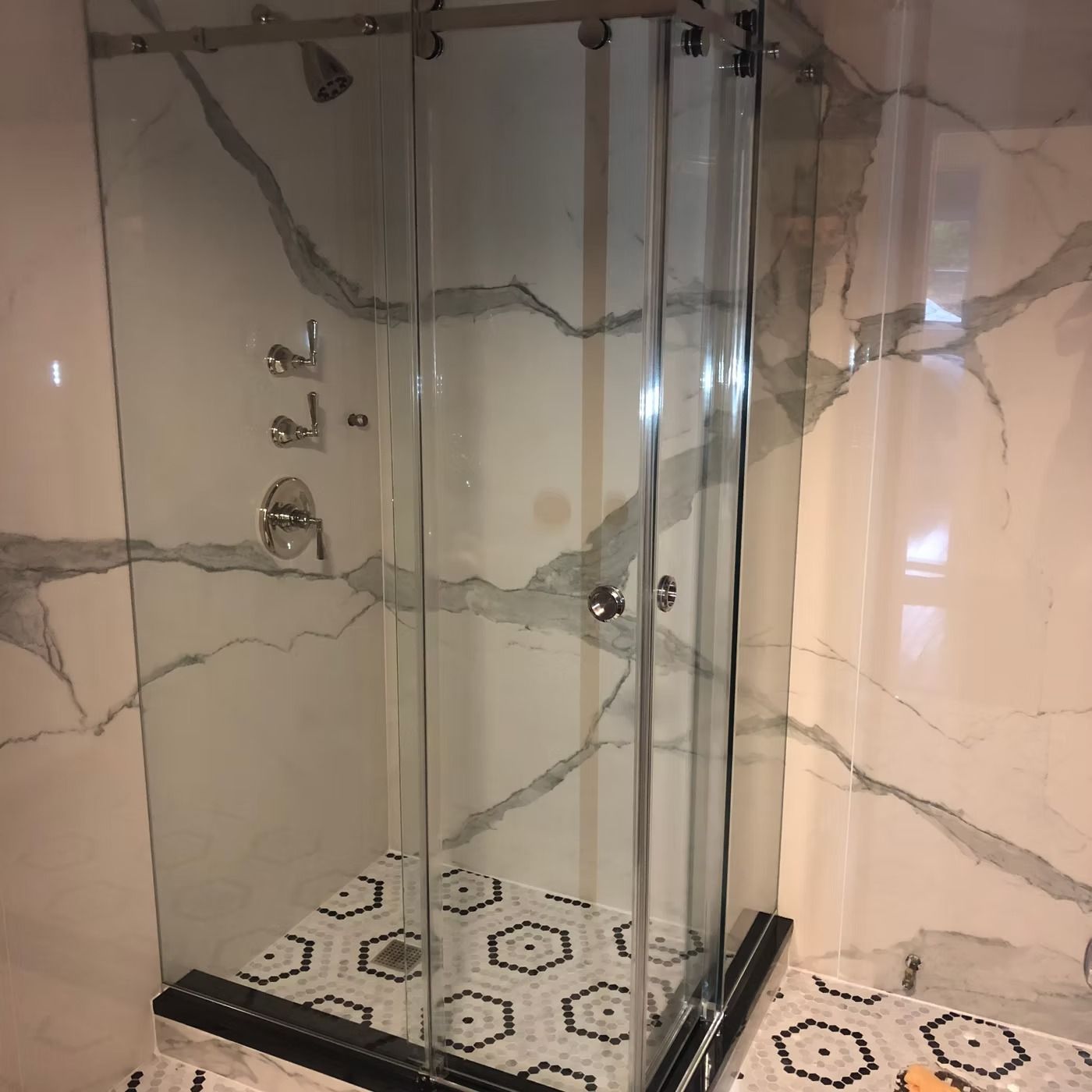 Glass-enclosed shower with beige marble walls and black-and-white hexagon tile floor.