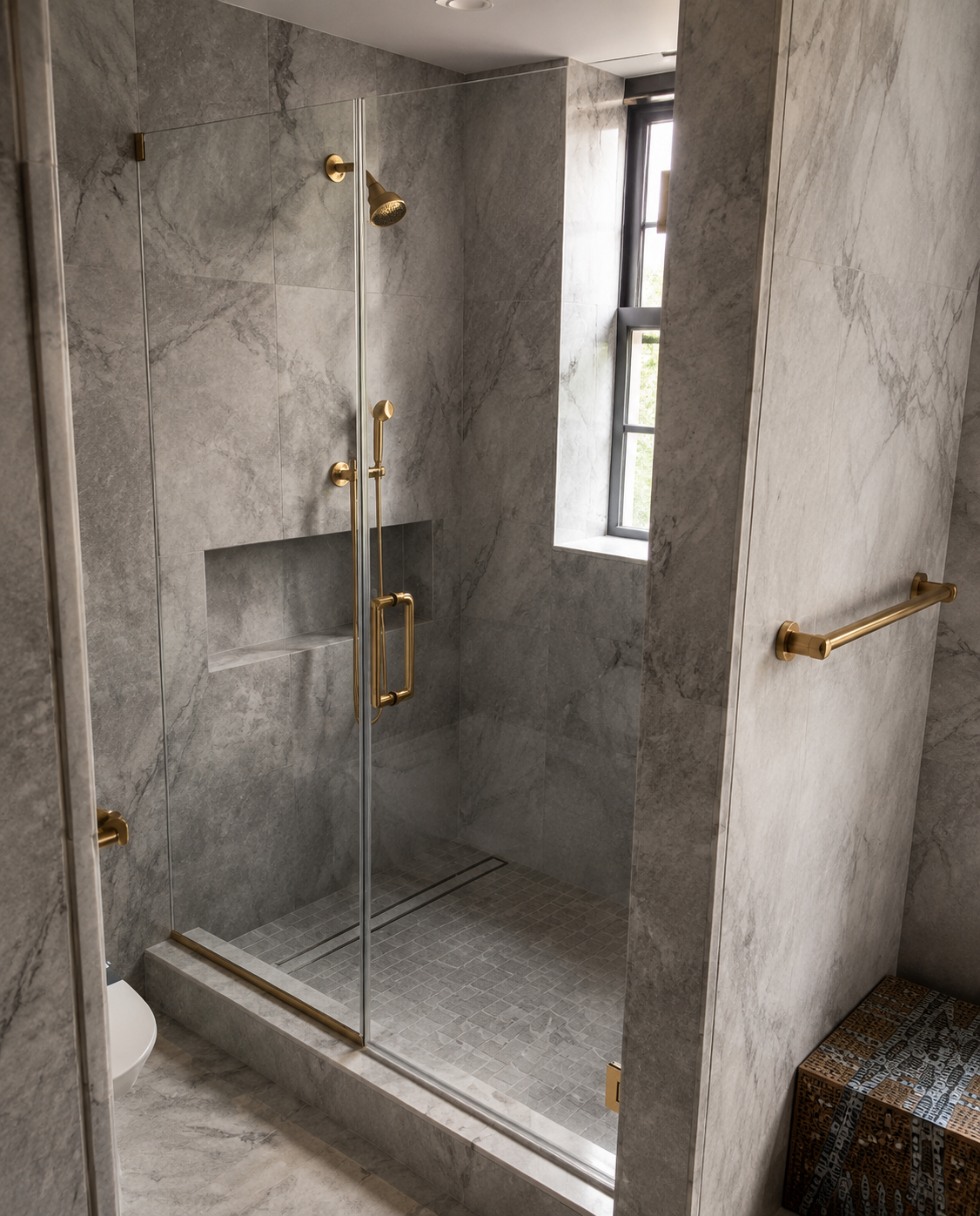 Glass shower enclosure in a gray tiled bathroom with gold fixtures and a small window