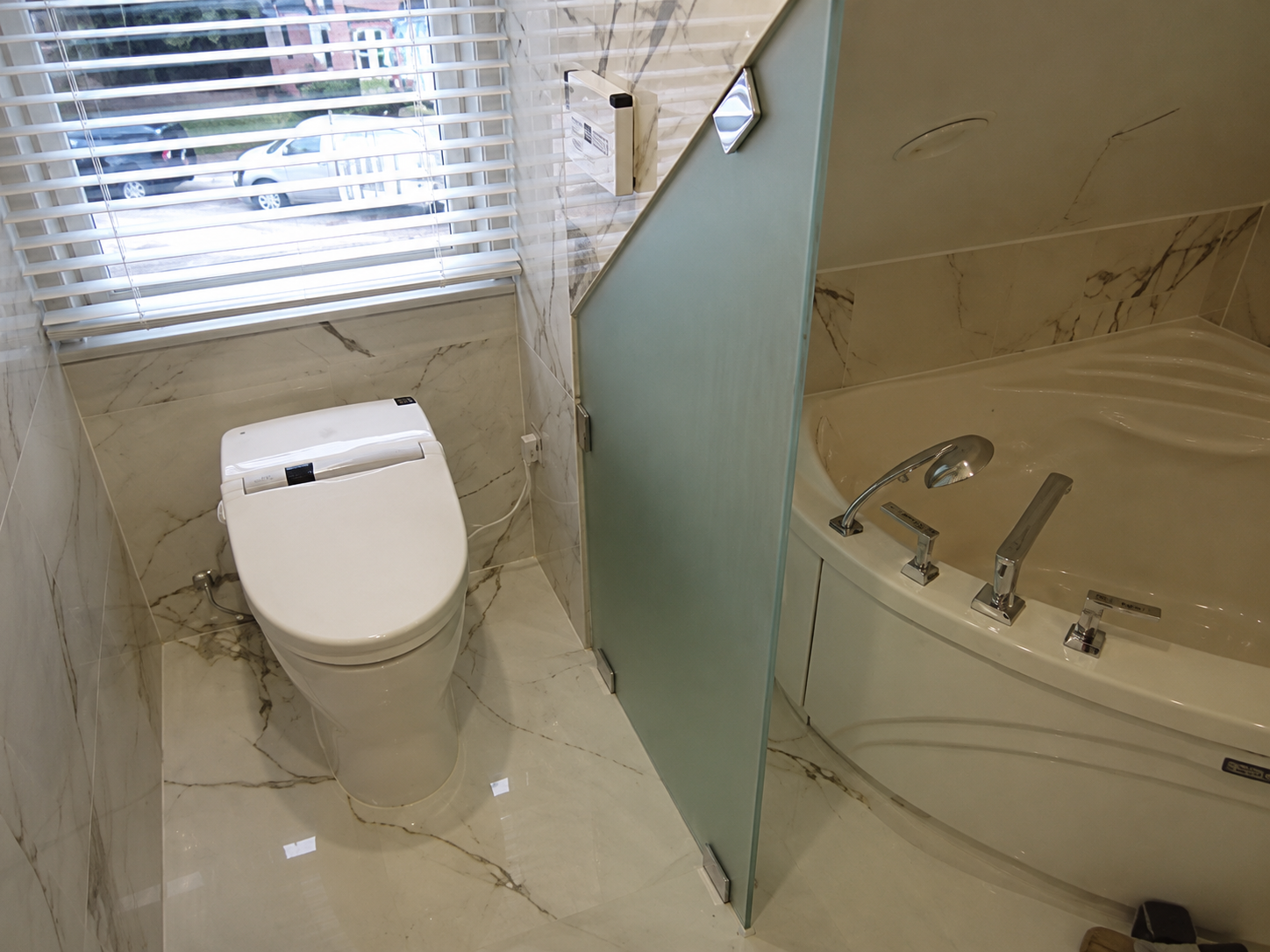 Small bathroom with toilet, glass shower partition, and corner bathtub under a window with blinds.