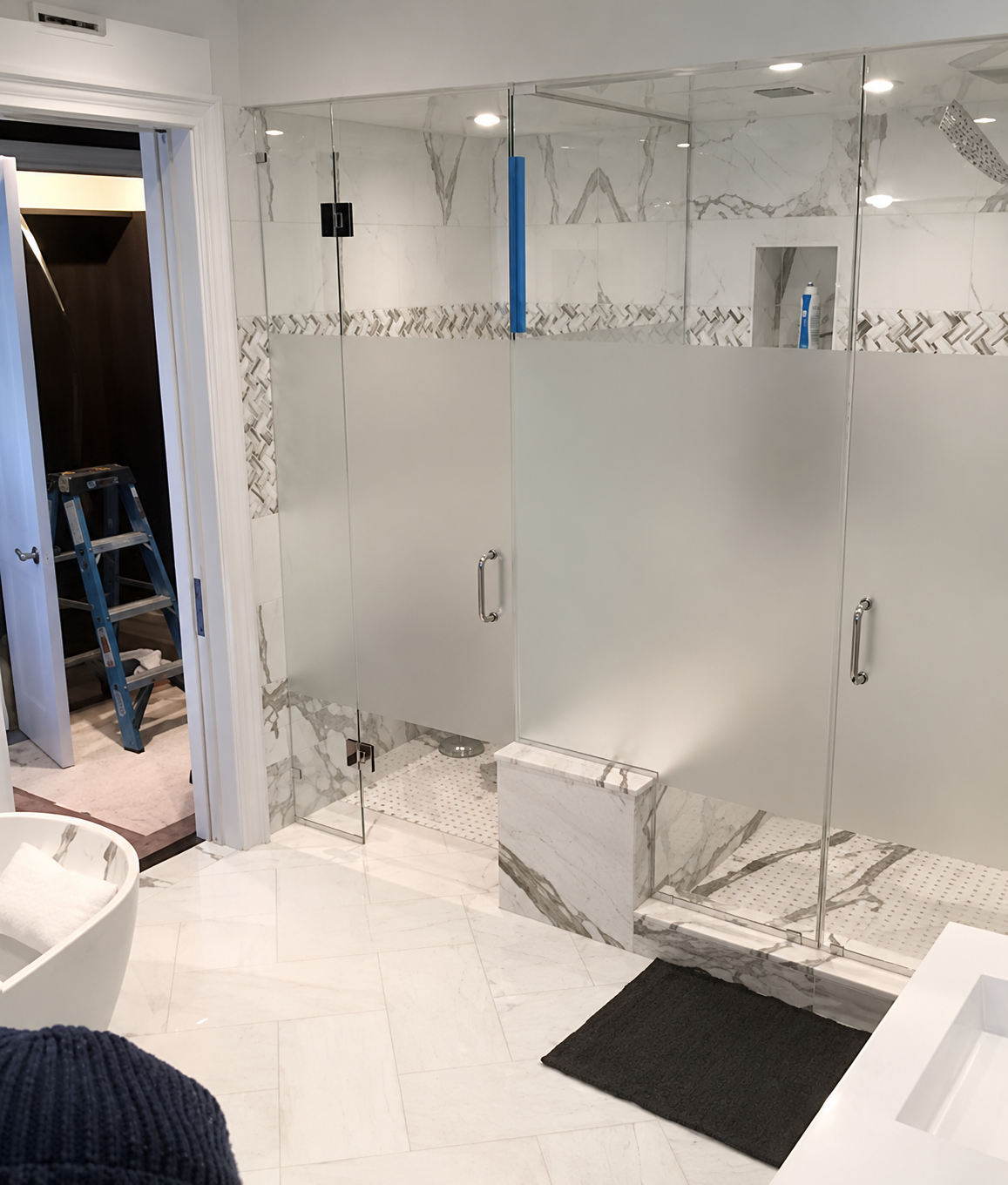 Modern bathroom with frosted glass shower doors, white tile walls, and a dark floor mat.