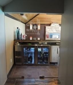 Small indoor bar area with wood ceiling, counter, glass-front mini fridge, and shelves of bottles and cups